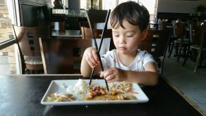 cute young chinese and caucasian boy learning to use chopsticks at restaurant